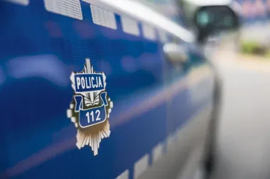 Fragment błyszczącego, niebieskiego policyjnego radiowozu z widocznym emblematem Policji i numerem 112, zaparkowanego przy drodze.